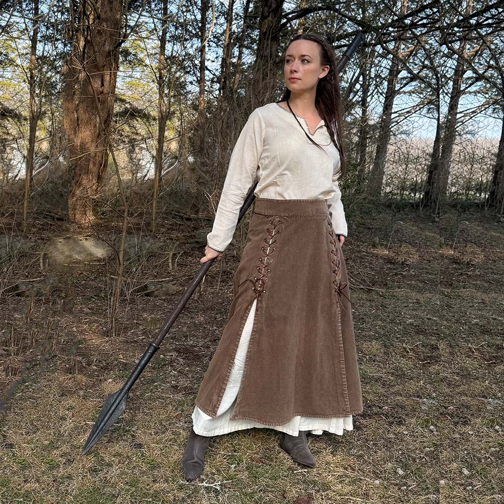 Wearing the Medieval Skirt- Ladies with a cream blouse, a woman stands in a sunlit wooded area holding a large spear, evoking the spirit of a historical reenactment as she gazes determinedly to the side.