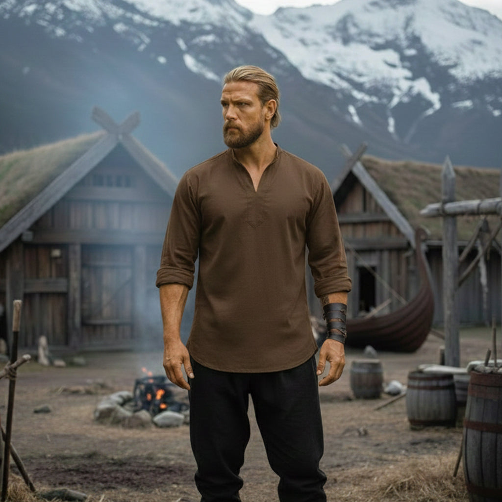 A man with long blonde hair and a beard stands before Viking-style buildings and snowy mountains, wearing the Warrior Shirt – Medieval Strength & Style- Unisex in brown and black pants, for an iconic Norse costume look. Fire burns behind him.