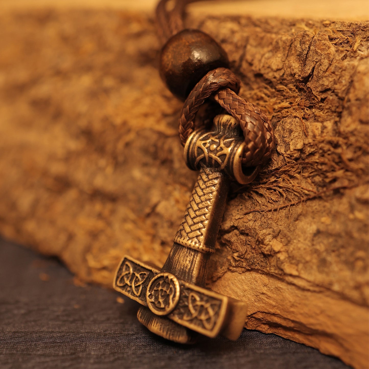 Close-up of the Norse Alley Thor’s Hammer Pendant in bronze with intricate carvings, displayed on a braided leather cord against textured wood—a bold tribute to Norse mythology jewelry.