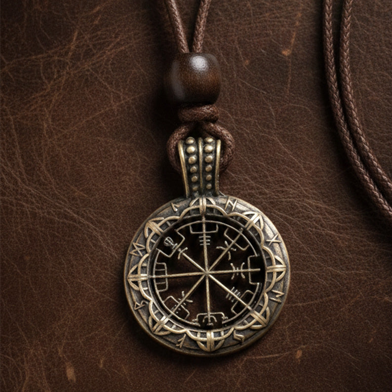 A round brass pendant with runic symbols and a compass-like design hangs from a brown cord on a textured leather surface.