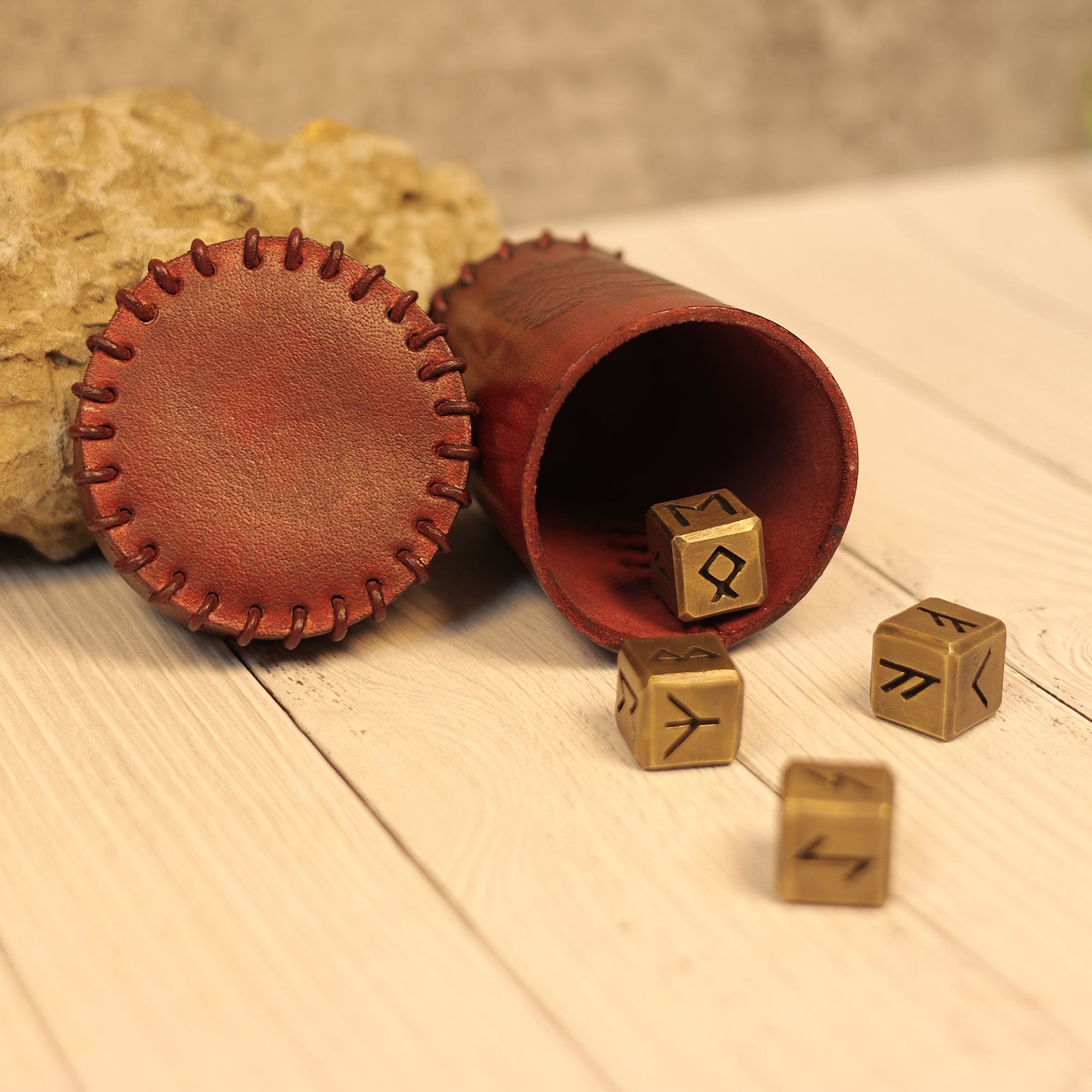 The Handcrafted Dark Red Leather Dice Cup with a stitched lid rests on a light wooden surface. Nearby, four rune-marked dice and a rough stone create an ideal scene for tabletop RPG accessories.