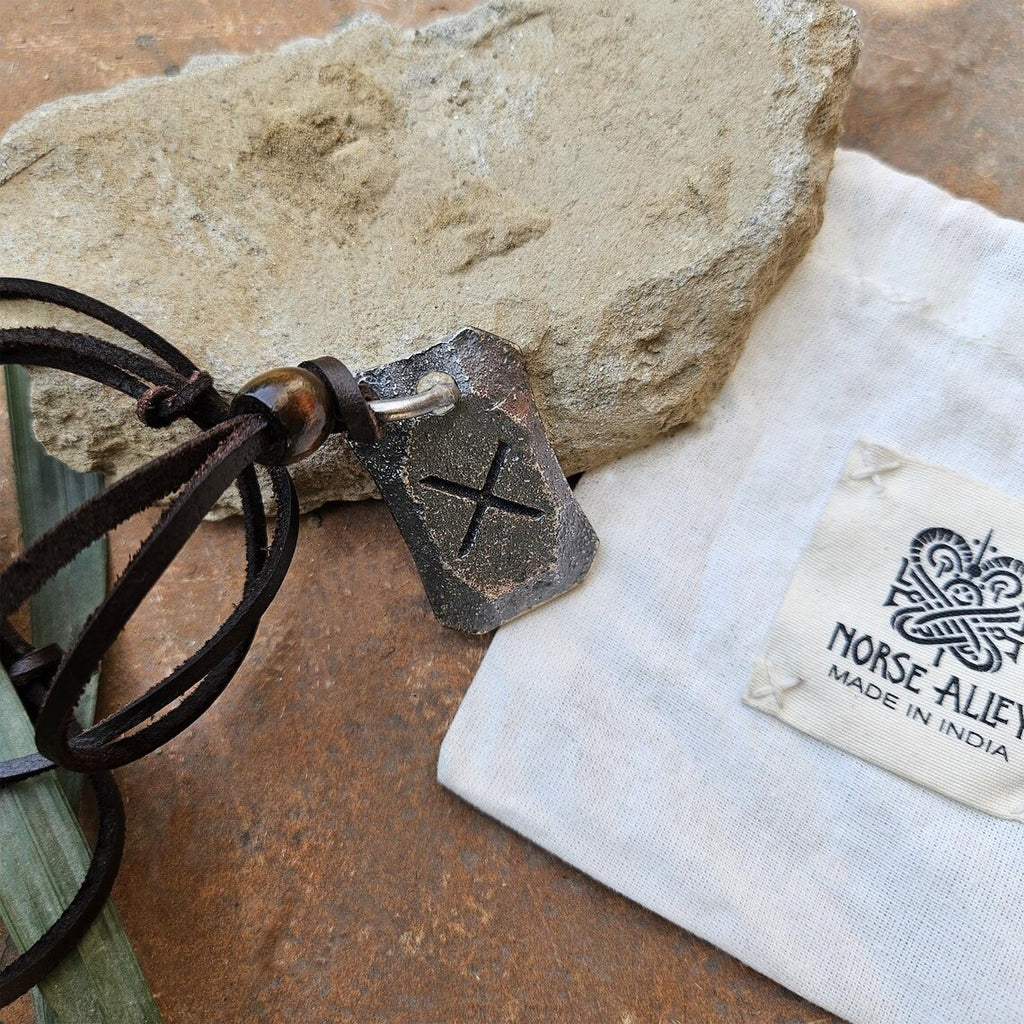 The Viking Rune Pendant Gebo features an engraved X Gebo rune on a leather cord, displayed on a stone. Next to it is a white pouch labeled Norse Aletheia, Made in India, enhancing its Norse mythology-inspired appeal.
