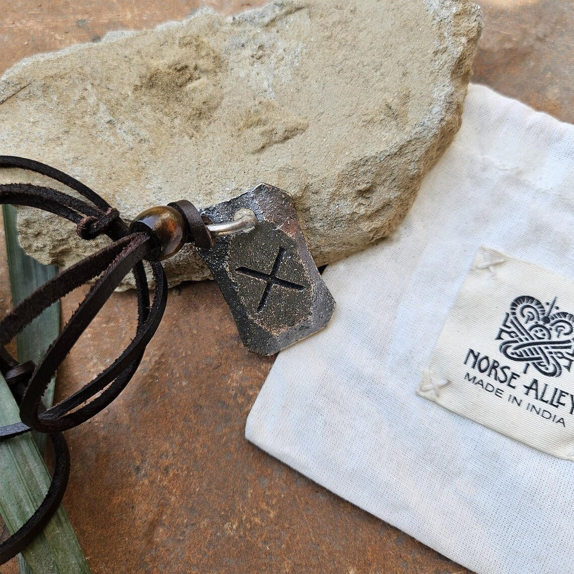 The Viking Rune Pendant Gebo features an engraved X Gebo rune on a leather cord, displayed on a stone. Next to it is a white pouch labeled Norse Aletheia, Made in India, enhancing its Norse mythology-inspired appeal.