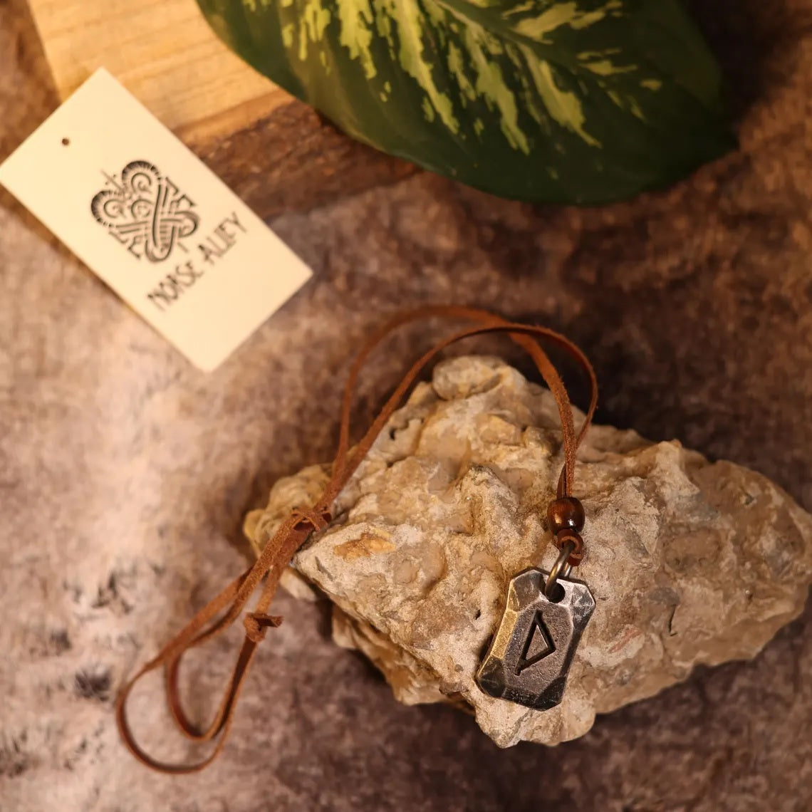 The Viking Rune Pendant Thurisaz, featuring a triangular rune on a leather cord, is shown atop rough stone next to greenery and a Moss Alley card with an intricate logo.