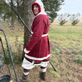 A person wearing the Viking Wool Coat for men, styled like a Norse klappenrock with a red-and-white hooded tunic, white trousers, and boots stands on grass holding a spear, with trees and an open field in the background.