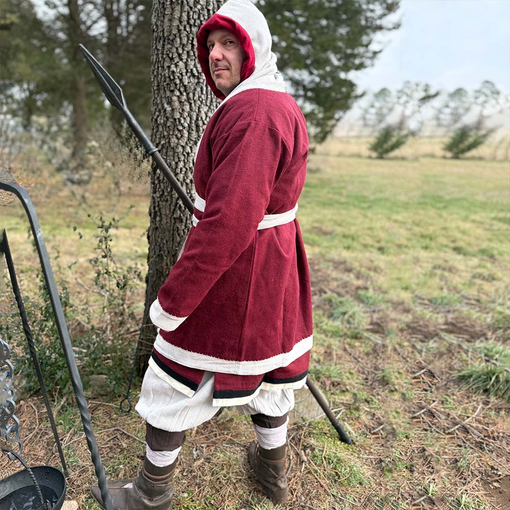 A person wearing the Viking Wool Coat for men, styled like a Norse klappenrock with a red-and-white hooded tunic, white trousers, and boots stands on grass holding a spear, with trees and an open field in the background.