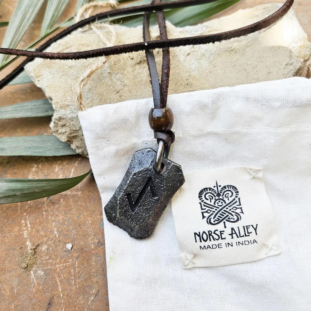 The Viking Rune Pendant Eihwaz, a dark metal piece on a leather cord, rests on a white Norse Alley Made in India pouch, with a green leaf and stone nearby.