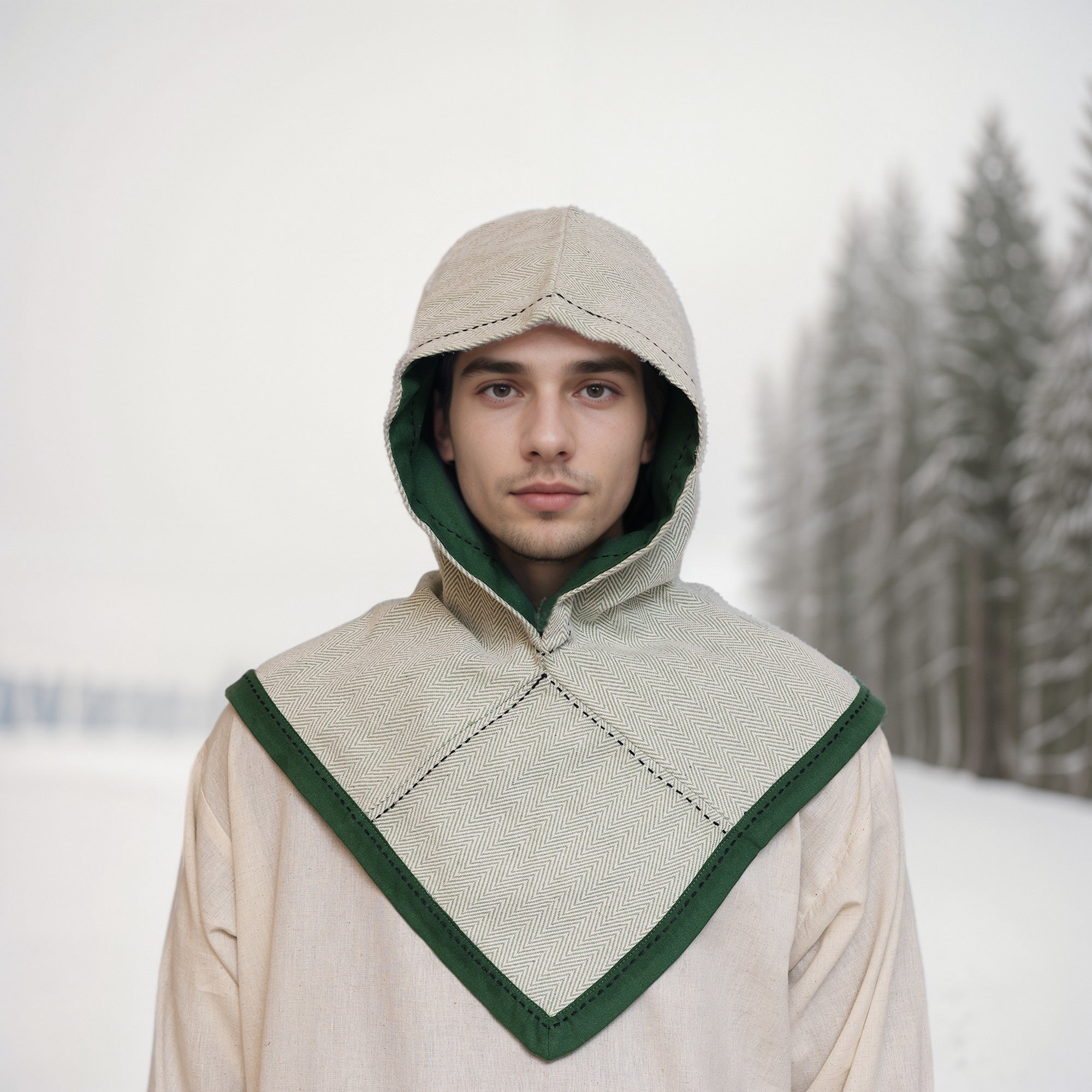 A young person wears a beige and green Reversible Viking Hood, Skjoldehamn, outdoors in a snowy landscape with pine trees—ideal for medieval reenactment.