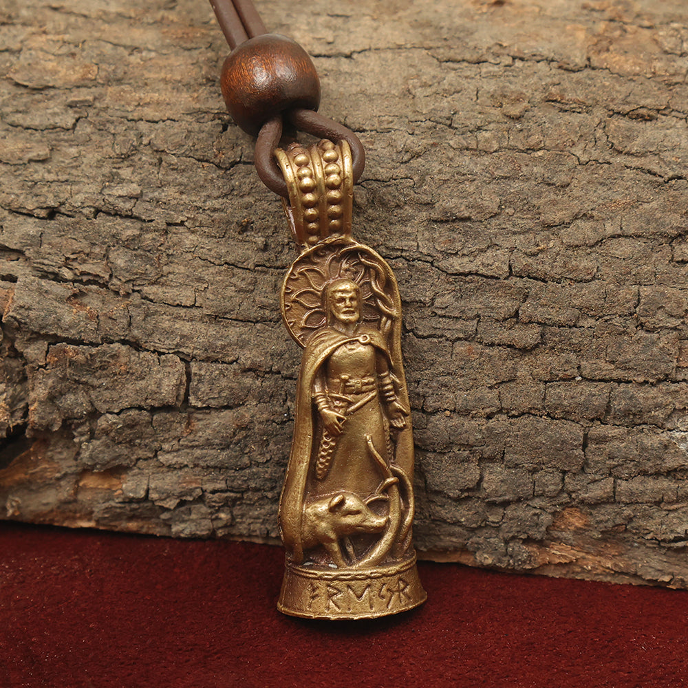 The Freyr Pendant – Lord of Abundance is a bronze Norse jewelry piece on leather cord, featuring a robed figure with halo above a coiled serpent or dragon. It rests on tree bark and red fabric; runic inscriptions in solid brass decorate the base.