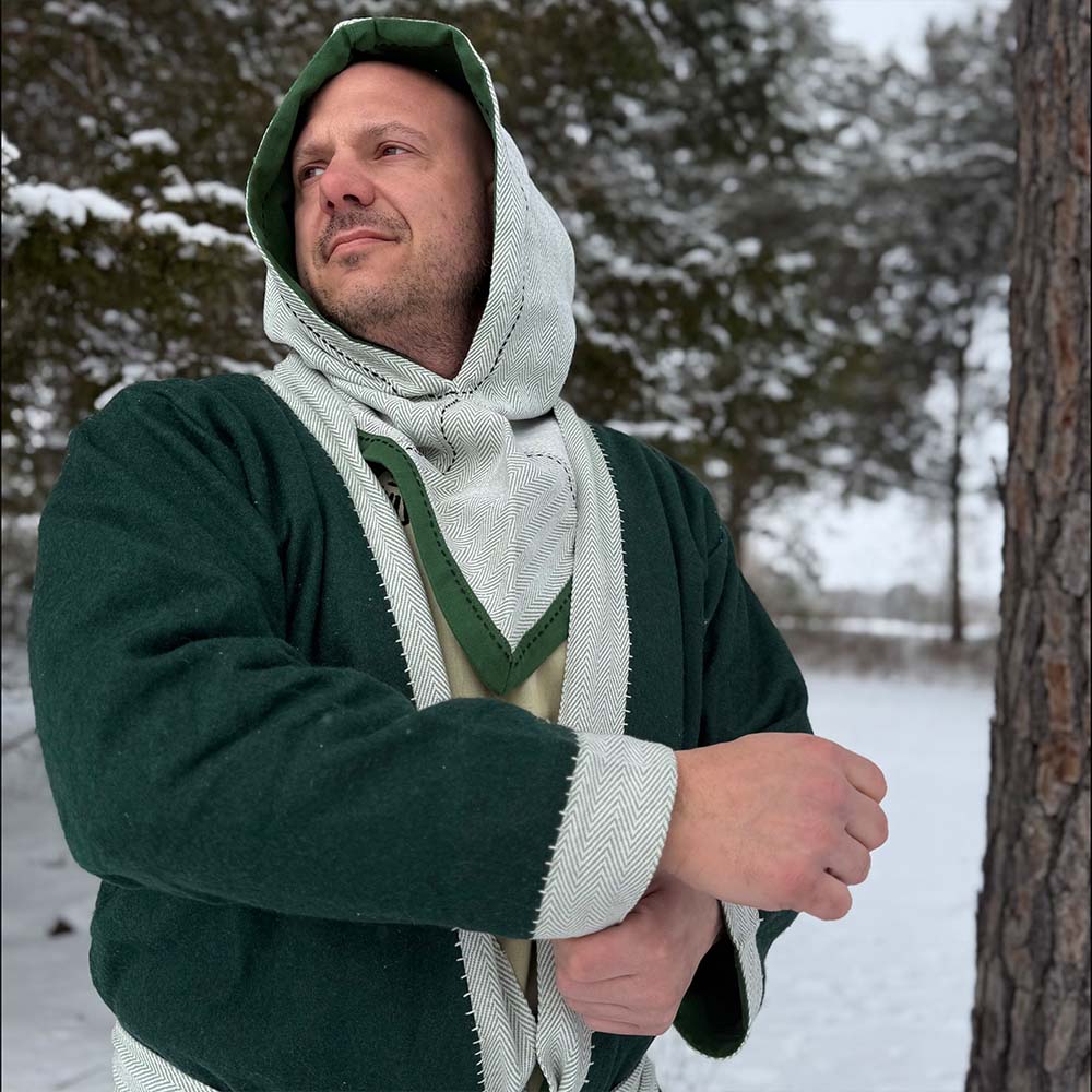 A man wearing a green Viking Wool Coat-Men's and a beige scarf stands in a snowy forest, looking to the side with one arm raised. Snow covers the ground and trees can be seen in the background.