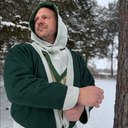 A man wearing a green Viking Wool Coat-Men's and a beige scarf stands in a snowy forest, looking to the side with one arm raised. Snow covers the ground and trees can be seen in the background.