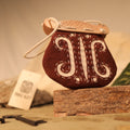 The "Gokstad" Coin Pouch, a small brown leather pouch with white embroidery and a wooden clasp, rests on bark with blurred objects and a card in the background.