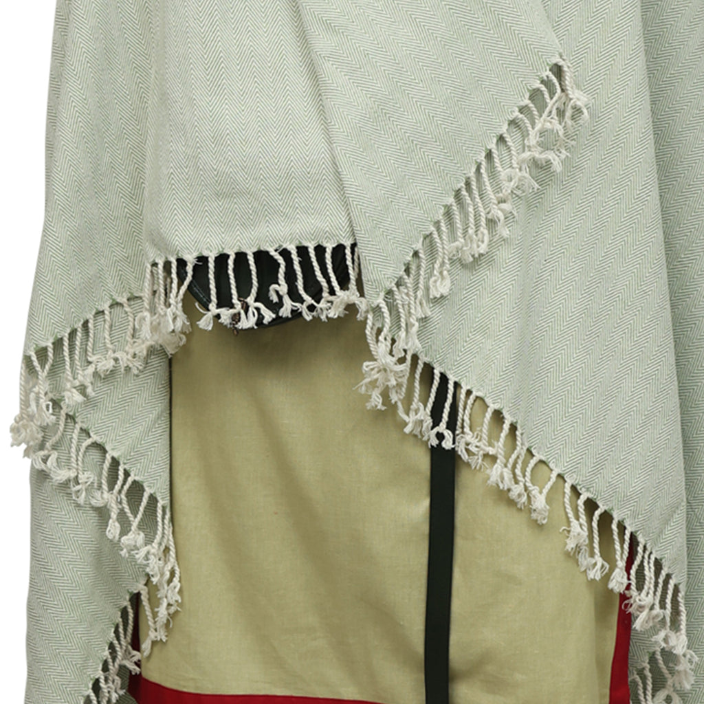 A close-up of Odin's Viking Cape- Unisex, showcasing its light green woven fabric with fringed edges, draped over a beige fabric featuring a green and red border. The textures and patterns of the cape are clearly visible.