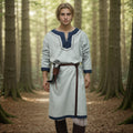 Wearing the Nordic/Viking Heritage Tunic for men in light blue with dark blue trim, dark pants, and a leather belt with pouch, he stands on a forest path amidst tall trees—a timeless renaissance ensemble.