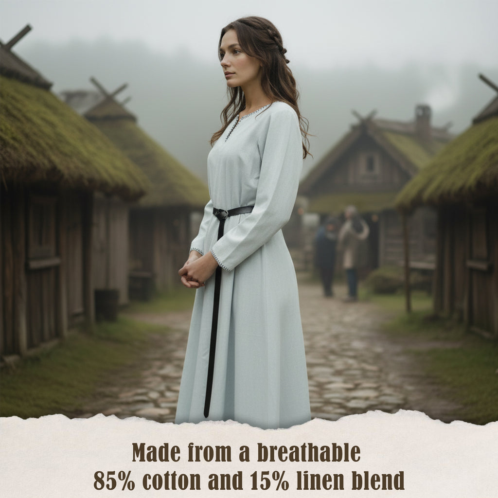 A woman in a pale blue Viking Thorndale Dress- Ladies stands on a cobblestone path in a rustic village. Text at the bottom: Crafted from breathable 85% cotton and 15% linen blend.