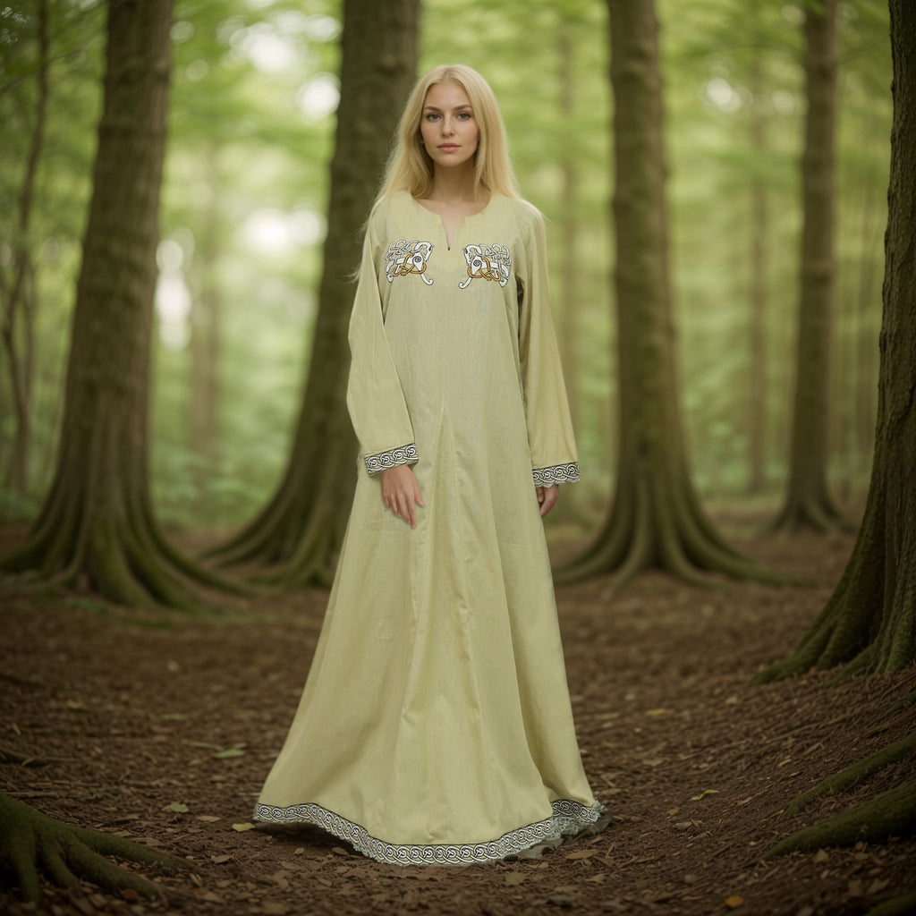 A woman with long blonde hair wears the Sleipnir Viking Dress for ladies, featuring embroidered patterns and light cotton-linen fabric, while standing in a sunlit forest surrounded by tall trees and lush greenery.