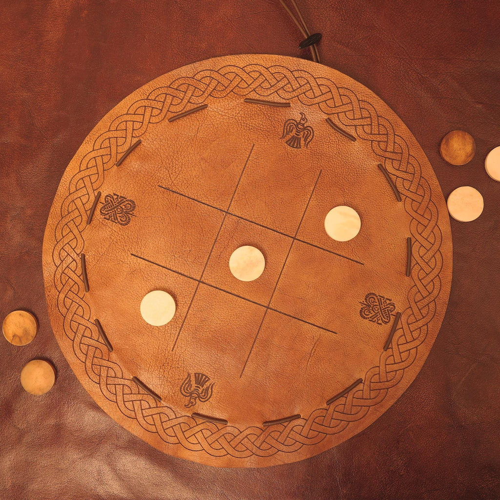 The Viking Leather Tic Tac Toe Game is a round, handcrafted board with Celtic knot designs and carved symbols, featuring light wooden pieces arranged on its brown leather surface.