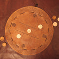 The Viking Leather Tic Tac Toe Game is a round, handcrafted board with Celtic knot designs and carved symbols, featuring light wooden pieces arranged on its brown leather surface.