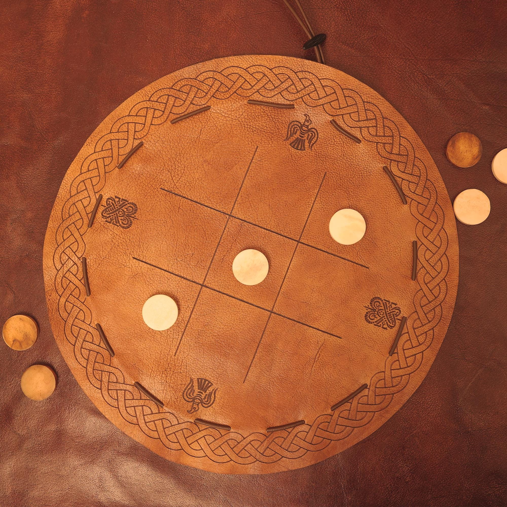 The Viking Leather Tic Tac Toe Game is a round, handcrafted board with Celtic knot designs and carved symbols, featuring light wooden pieces arranged on its brown leather surface.