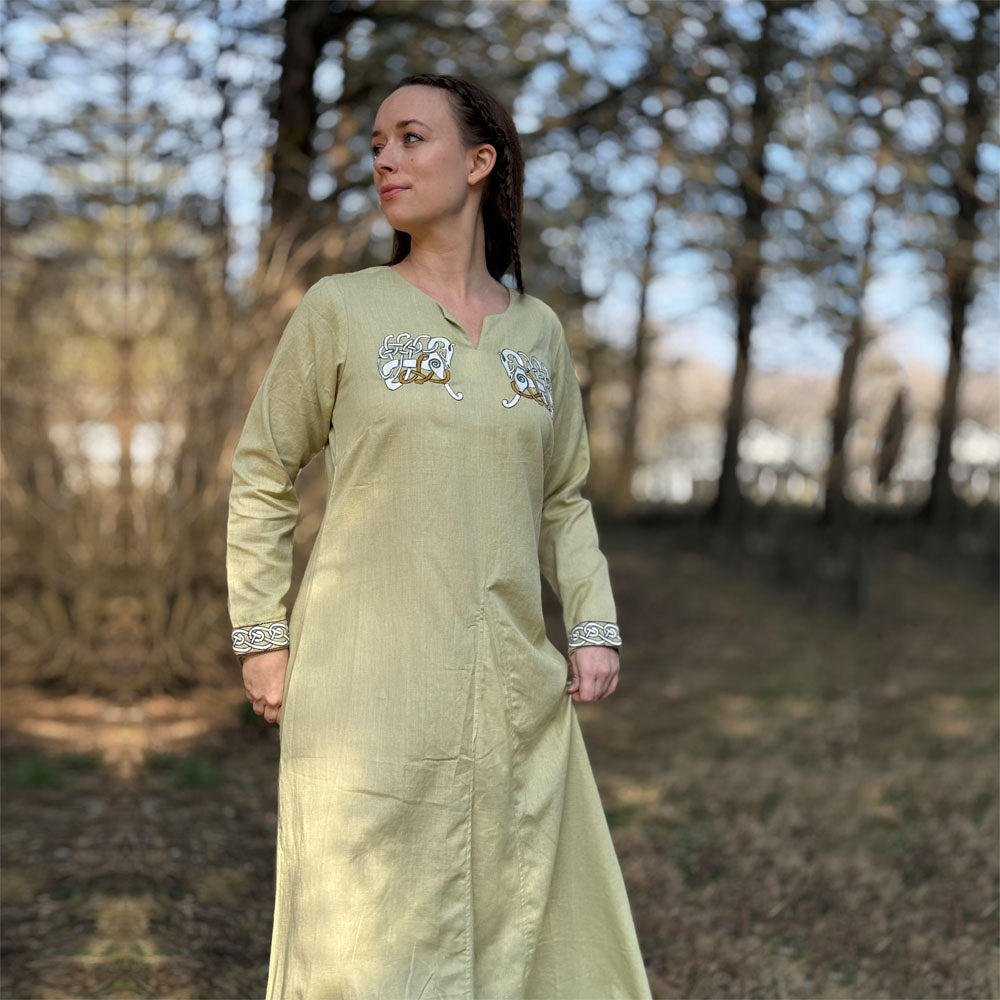 A woman wears the Sleipnir Viking Dress- Ladies in light yellow cotton-linen, standing outdoors in a sunlit wooded area with blurred trees and brown grass behind her. She looks to the side with her hands by her sides.