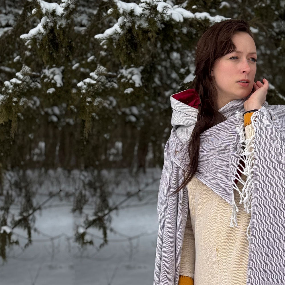 A woman stands before snow-covered evergreens, wearing Odin's Viking Cape–Unisex over light attire. She gently touches her face and gazes into the distance, embodying Norse spirit amid a wintry forest scene.