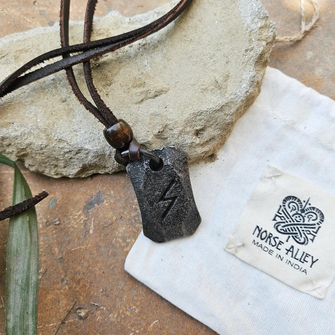 The Viking Rune Pendant Sowilo, featuring a lightning bolt symbol on a leather cord, rests on a rock beside a white Norse Alley bag marked "Made in India," drawing inspiration from Norse mythology.