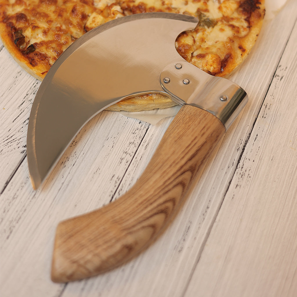 A Pizza Axe with a brown scabbard and stainless steel blade rests on a pizza atop a white wooden surface.