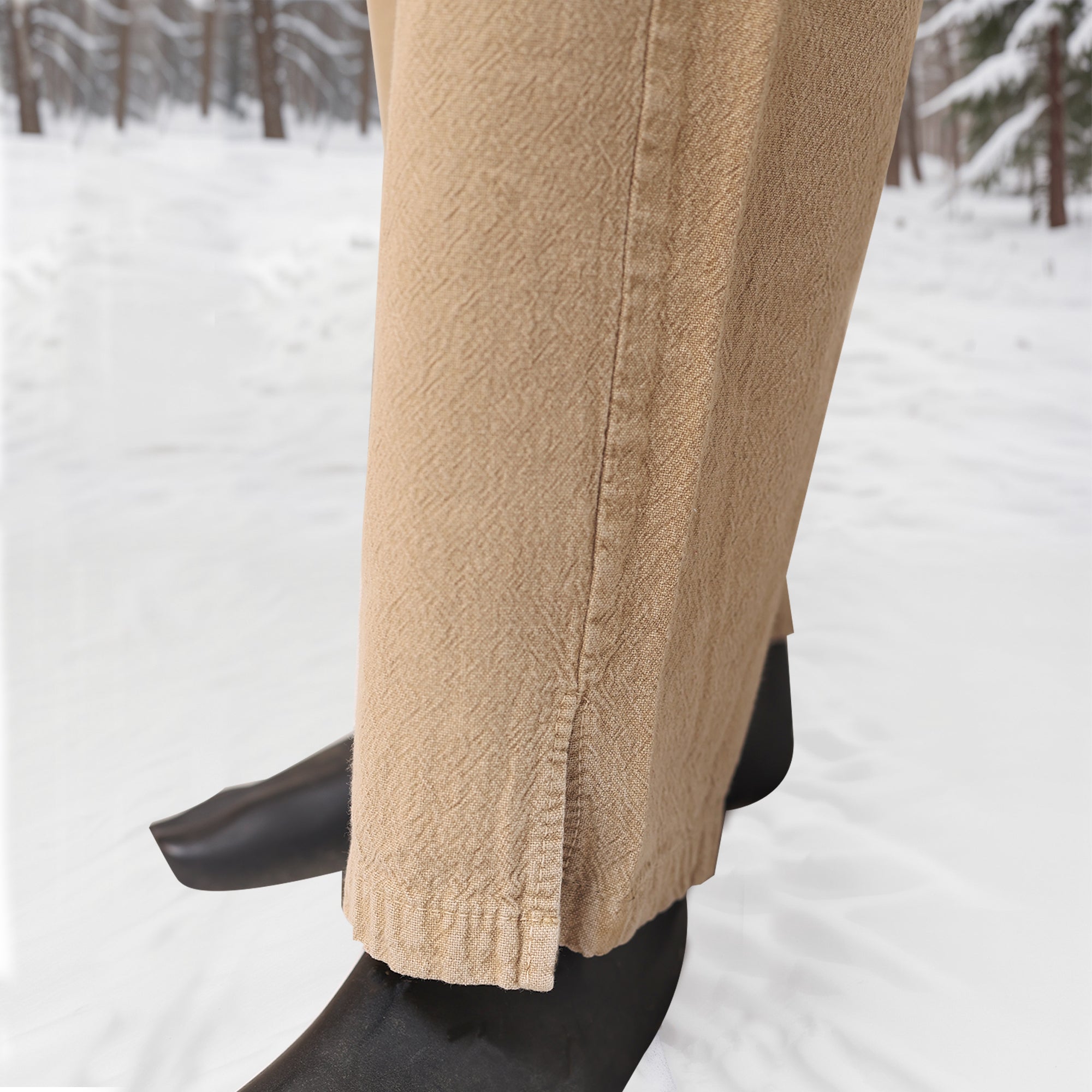 A close-up shows a person wearing Warrior Pants - Men's with black shoes, standing on snowy ground among trees. These tan textured trousers feature a small hem slit, ideal for Norse reenactment or winter wear.