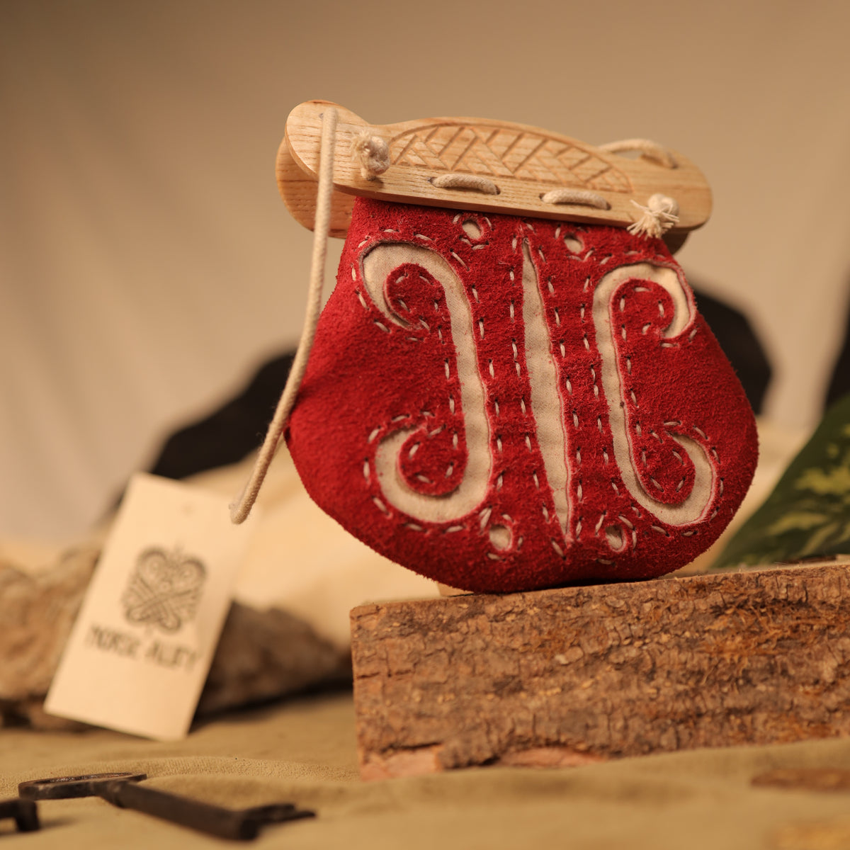 A red, embroidered leather "Gokstad" Coin Pouch with white stitching and wooden accents rests on wood. A tag featuring a Nordic-style logo and NORD ALSIT hints at its Norse bag inspiration in the background.