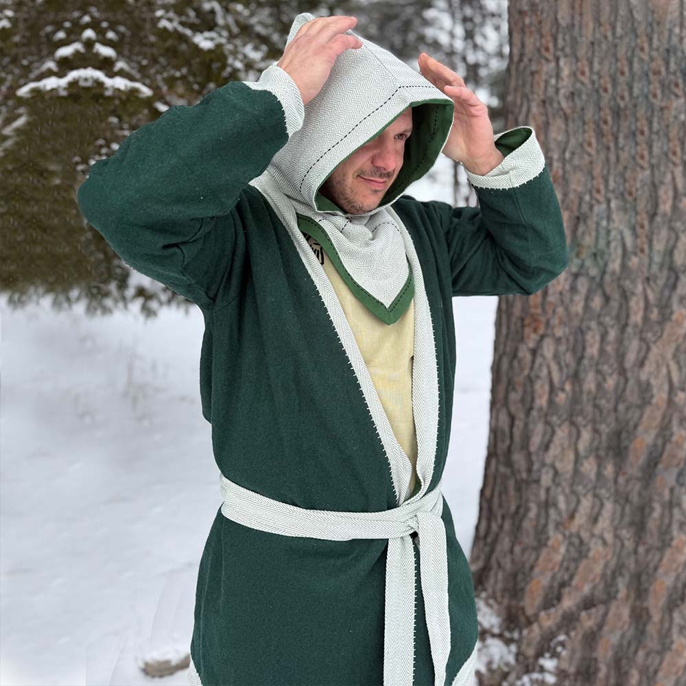 A person wearing a green and grey Reversible Viking Hood, Skjoldehamn, stands outside in the snow by a tree, adjusting their hood. Snow and evergreens surround them, evoking the atmosphere of a medieval reenactment.