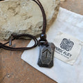 The Viking Rune Pendant Tiwaz, featuring an engraved arrow symbol, hangs from a brown leather cord and rests on a Norse Alley pouch labeled "Made in India," with a stone backdrop.