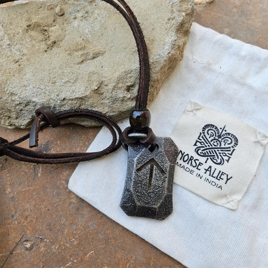 The Viking Rune Pendant Tiwaz, featuring an engraved arrow symbol, hangs from a brown leather cord and rests on a Norse Alley pouch labeled "Made in India," with a stone backdrop.