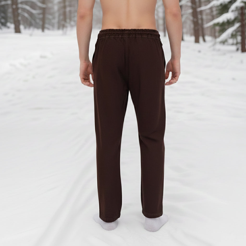 A shirtless man stands barefoot in the snow, seen from behind, wearing Warrior Pants- Men's. Snow-covered ground and trees are visible in the background.
