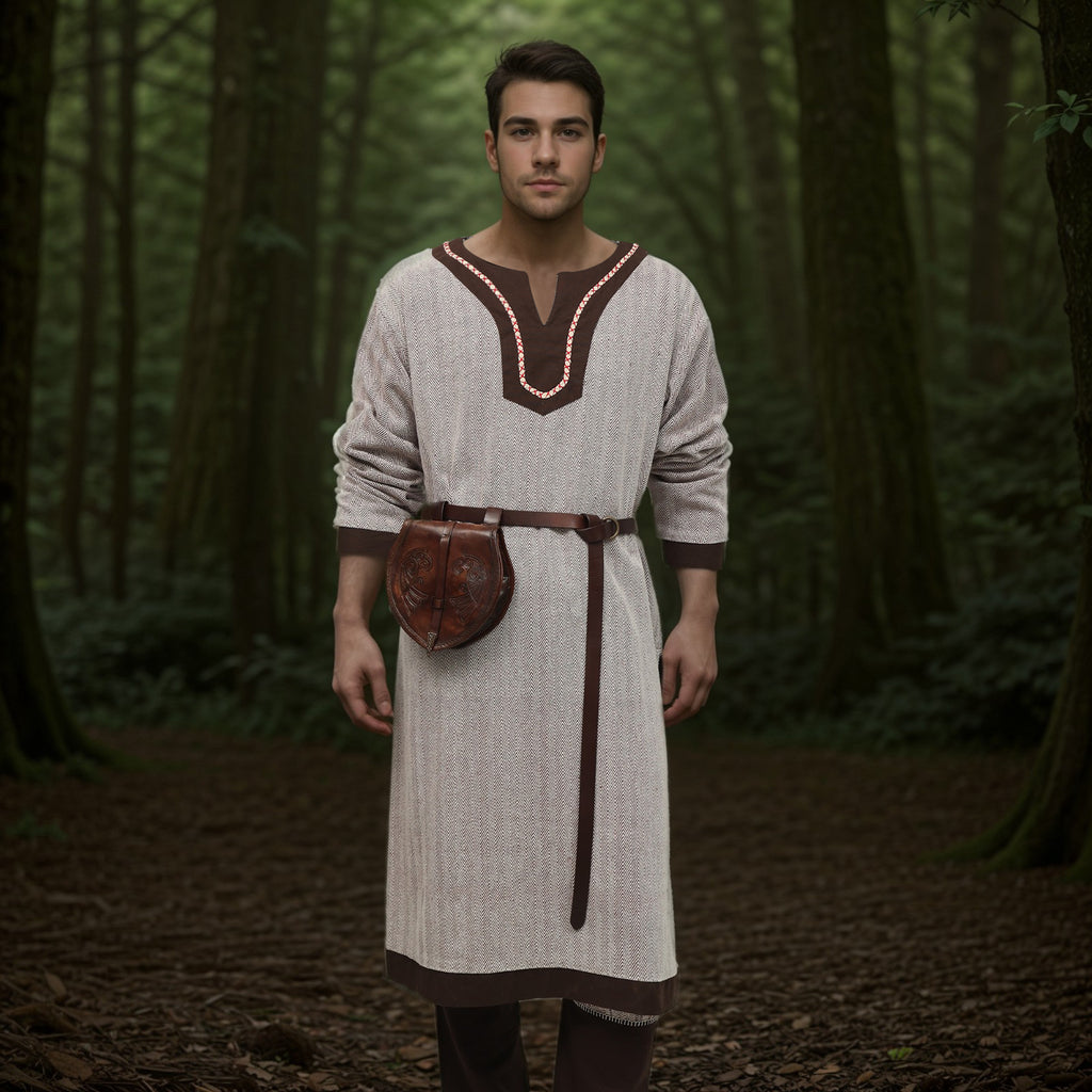 Wearing the Nordic Runeweave Viking Tunic for men, complete with a leather belt and pouch, a man stands facing the camera in a dense green forest surrounded by tall trees and lush foliage.