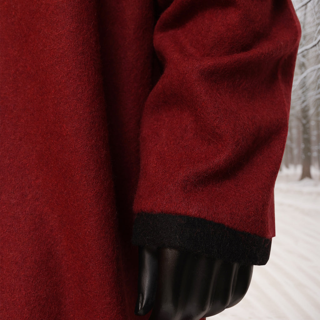 A close-up of a man wearing the Viking Woolen Tunic—Men's, a red wool coat with black-trimmed sleeve inspired by Viking tunic designs, standing outdoors in a snowy, tree-lined setting; only his arm and part of the tunic are visible.