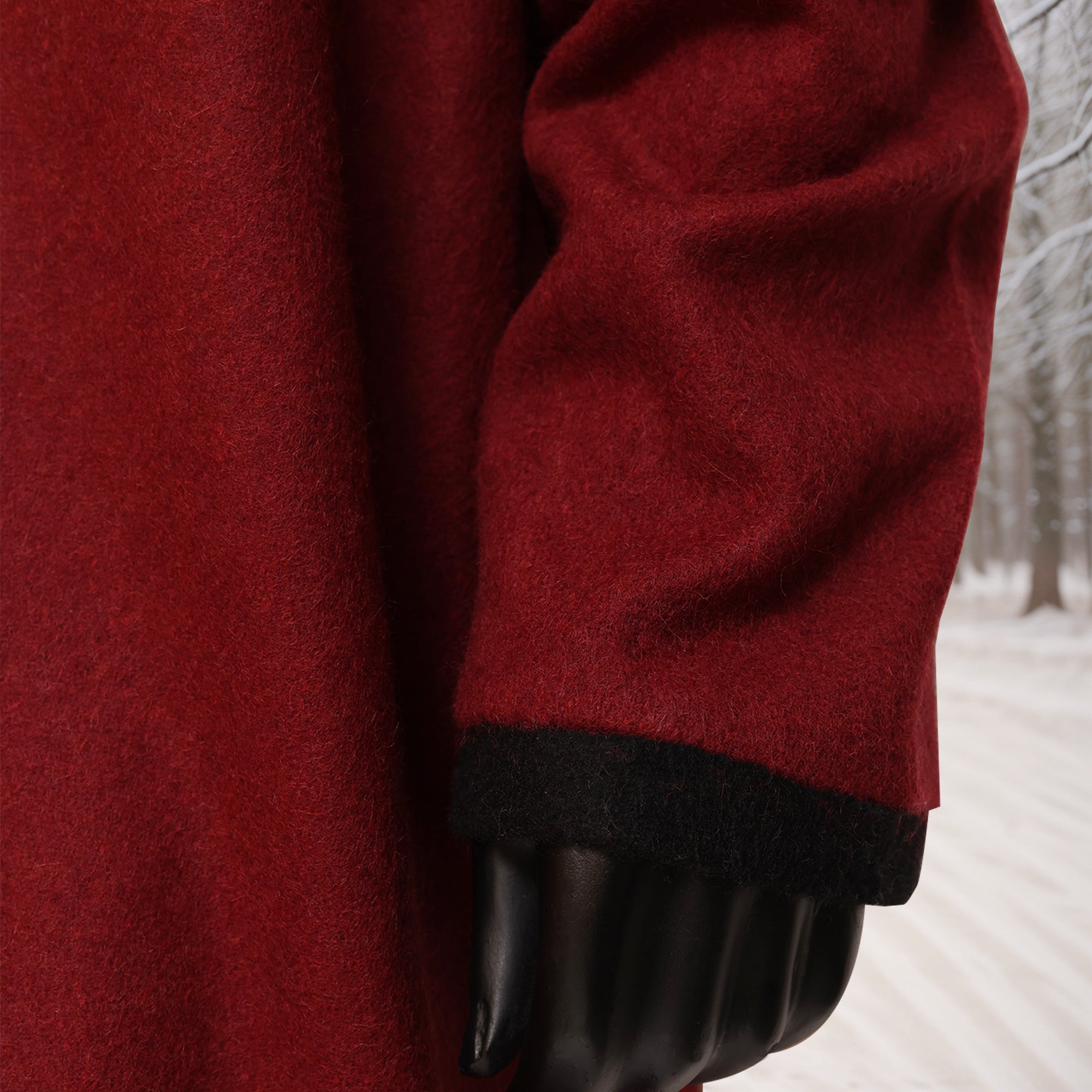 A close-up of a man wearing the Viking Woolen Tunic—Men's, a red wool coat with black-trimmed sleeve inspired by Viking tunic designs, standing outdoors in a snowy, tree-lined setting; only his arm and part of the tunic are visible.