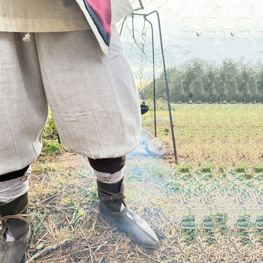 Wearing Nordic Nomad Viking Pants for men, a reenactor stands on grass by a campfire with a pot hanging above the flames and an open field in the background.