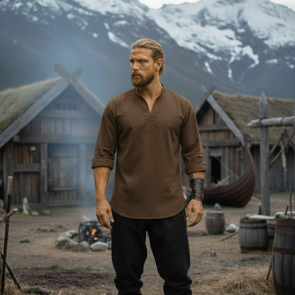 A man wearing the Warrior Shirt – Medieval Strength & Style – Unisex stands before wooden Viking houses, with a firepit, longship, barrels, and snowy mountains in the background.