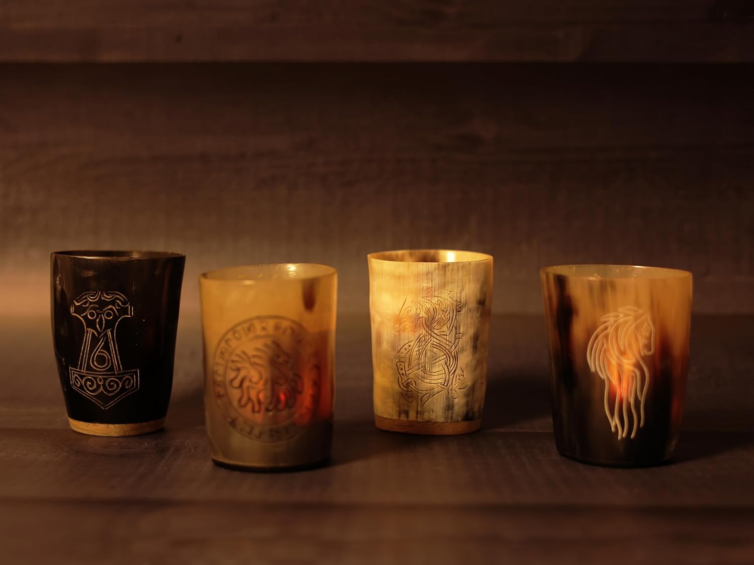 Four Norse Alley Viking Drinking Horn Shot Glasses, each with intricate Norse designs, are lined up on a dark wooden surface, warmly lit—ideal as unique drinkware or striking home decor accents.