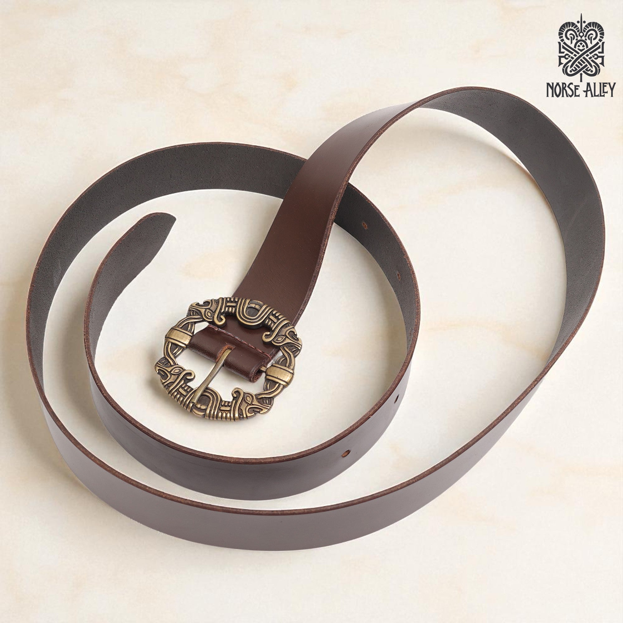 The Norse Alley The Drakkar Belt is a long brown Viking leather belt with an ornate Norse-patterned metal buckle, displayed on a light surface with the Norse Alley logo in the top right corner.