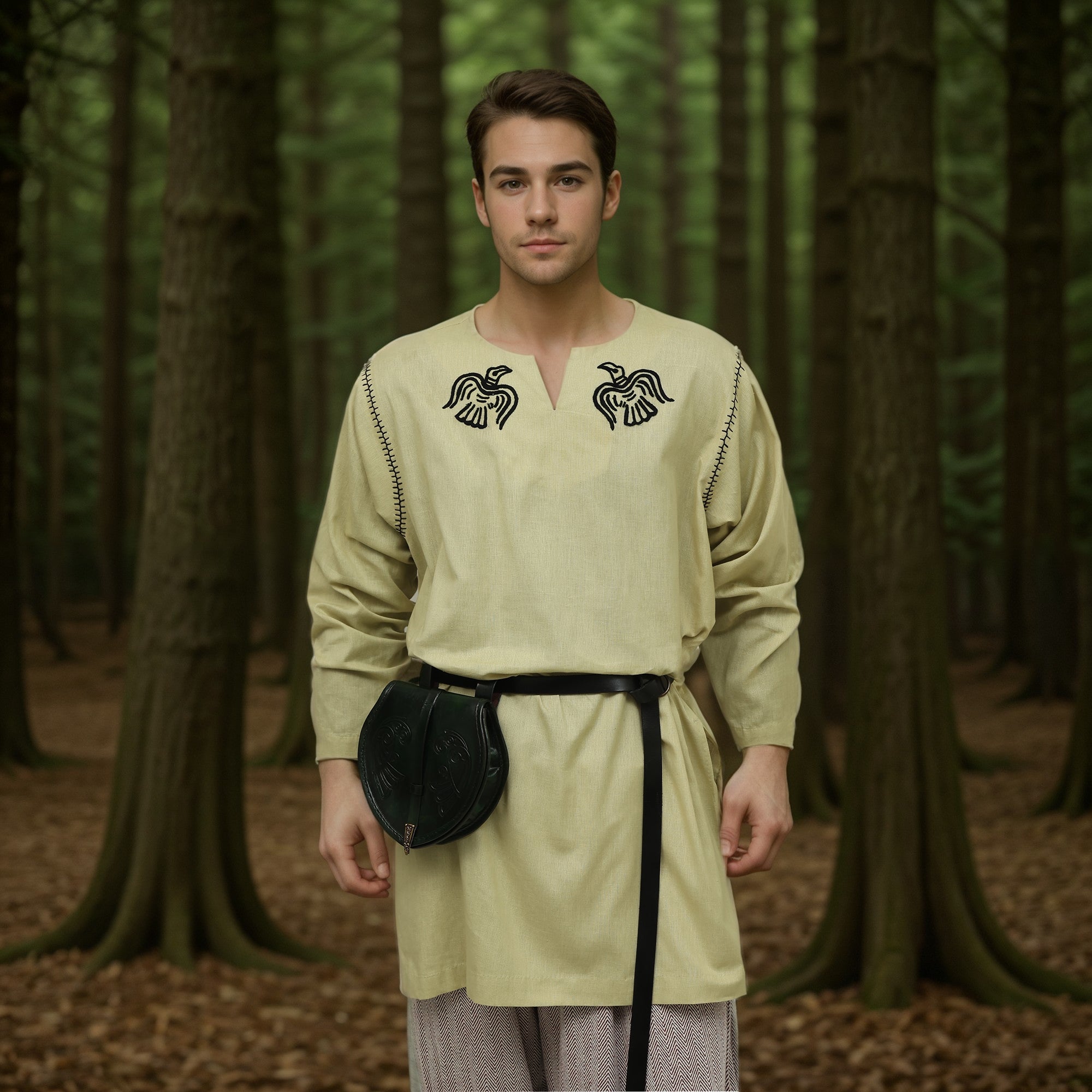A man stands in a forest of tall trees and fallen leaves, wearing the Odin's Raven Tunic—Men's, a beige Viking tunic featuring black raven embroidery and paired with a black belt.