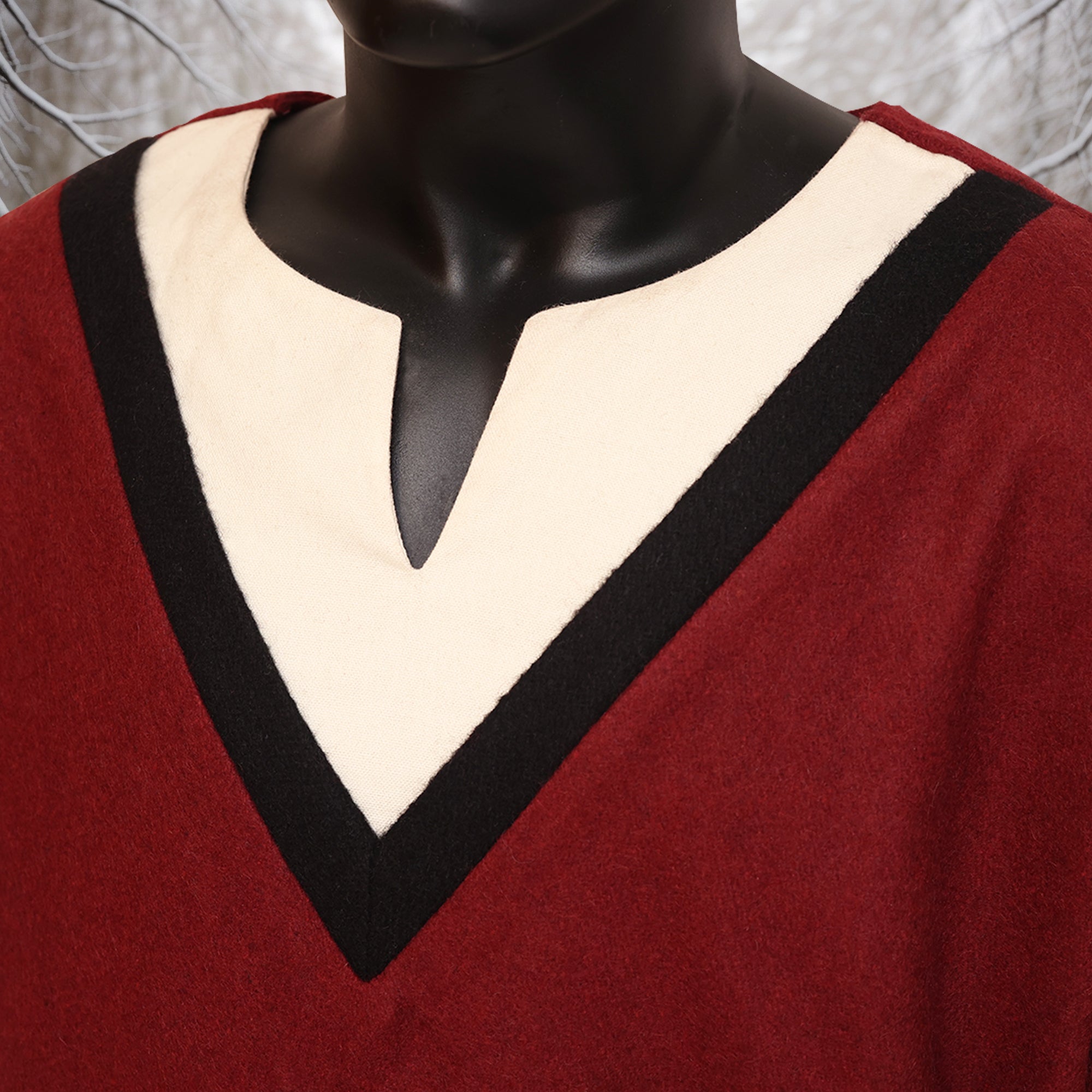 Close-up of a person wearing the Viking Woolen Tunic- Men's in red, featuring a cream and black V-shaped neckline. The face is not visible and the gray background is out of focus.