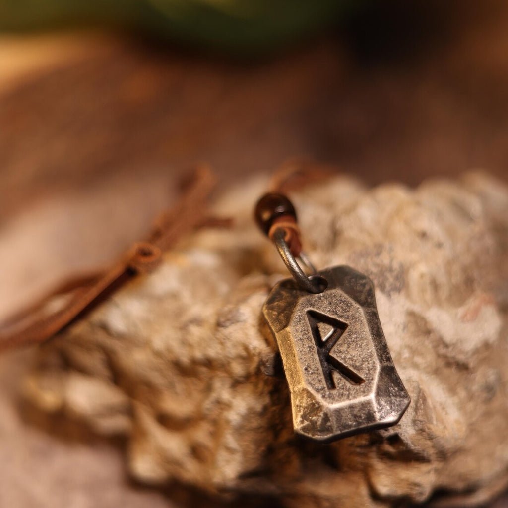 The Viking Rune Pendant Raido features a metallic Norse rune "R" engraved charm on a brown cord, displayed atop textured stone. The softly blurred background enhances its connection to Norse mythology.