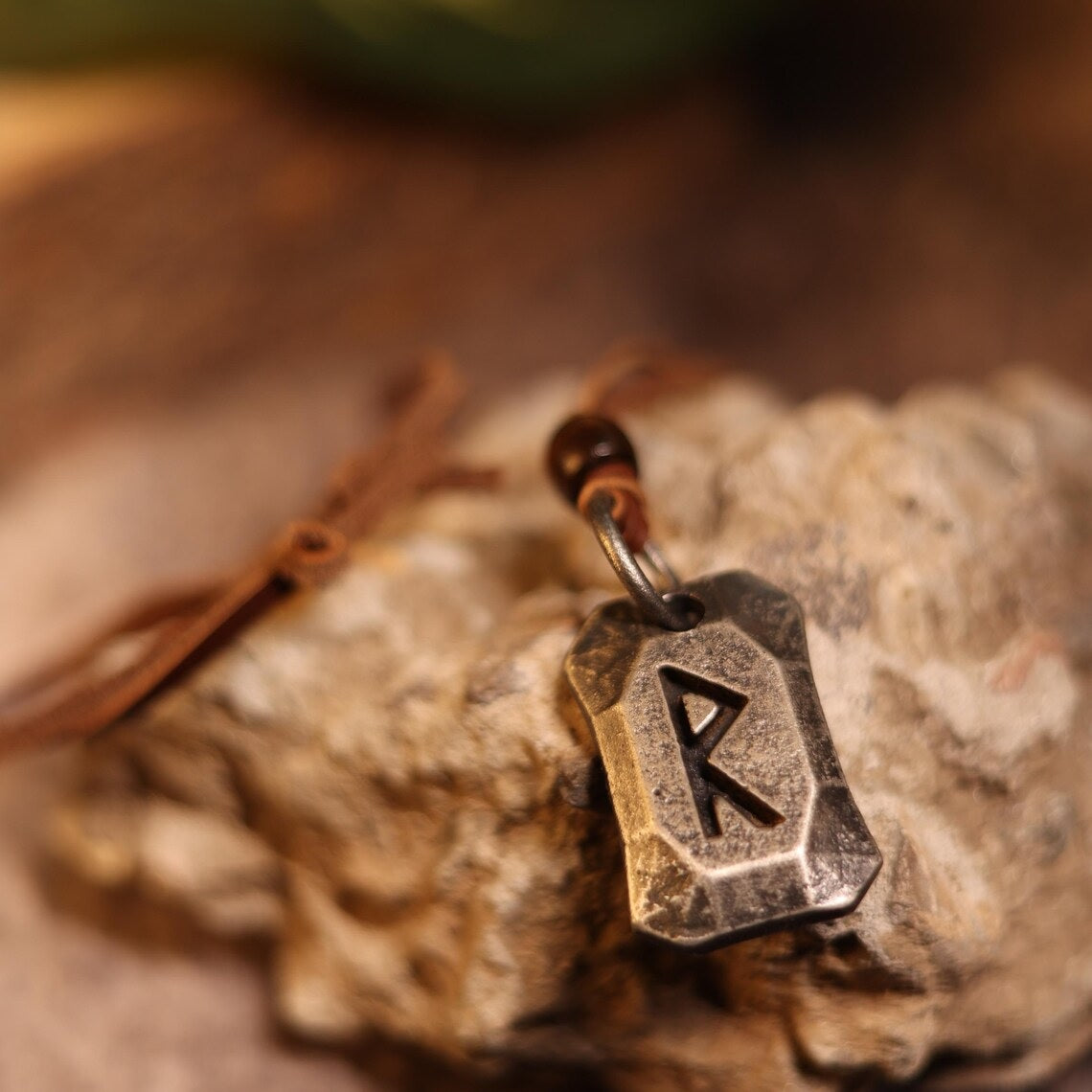 The Viking Rune Pendant Raido features a metallic Norse rune "R" engraved charm on a brown cord, displayed atop textured stone. The softly blurred background enhances its connection to Norse mythology.