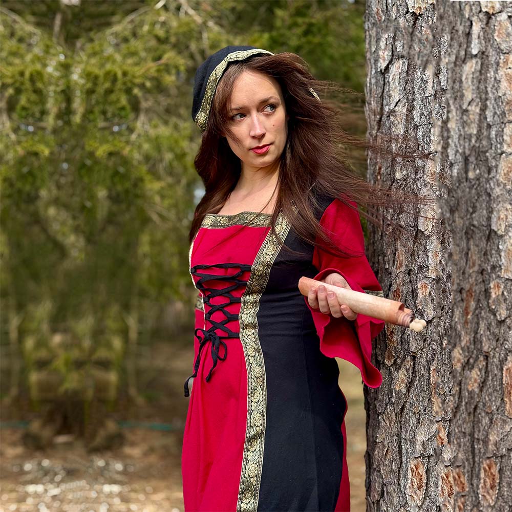 A woman wearing the Dark Majesty Medieval Dress- Ladies in red and black with gold trim stands by a tree, holding a rolling pin. Her loose hair blows in the wind, surrounded by trees and greenery.