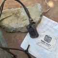 The Viking Rune Pendant Jera in black hangs on a leather cord next to a Norse Alley "Made in India" white cloth pouch, both set on rough stone.