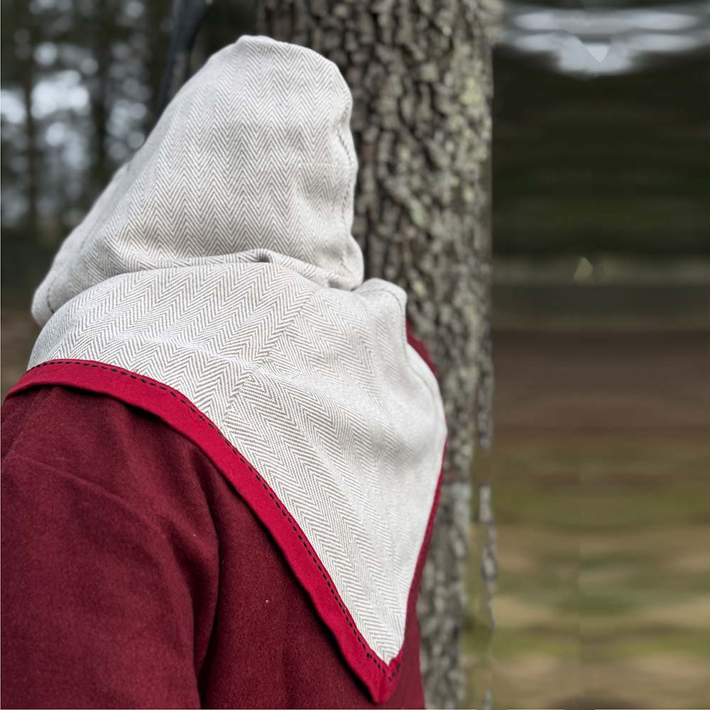 A person wearing the Reversible Viking Hood, Skjoldehamn with red trim over a red garment stands outdoors near trees, facing away from the camera. The blurred natural backdrop enhances the medieval reenactment feel.