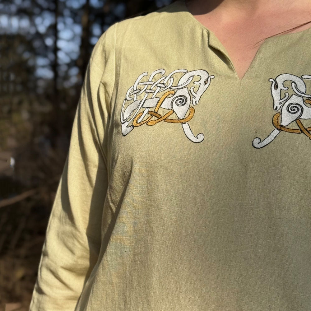 A person wears the Sleipnir Viking Dress—Ladies, a light yellow cotton-linen gown featuring embroidered Norse-inspired animal designs near the shoulders, standing outdoors with blurred trees in the background.
