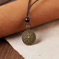 The Traveler’s Compass Pendant Necklace features a round antique bronze Viking design on a dark leather cord with a wooden bead, displayed on light fabric beside rolled tan leather—a striking piece of Norse jewelry.