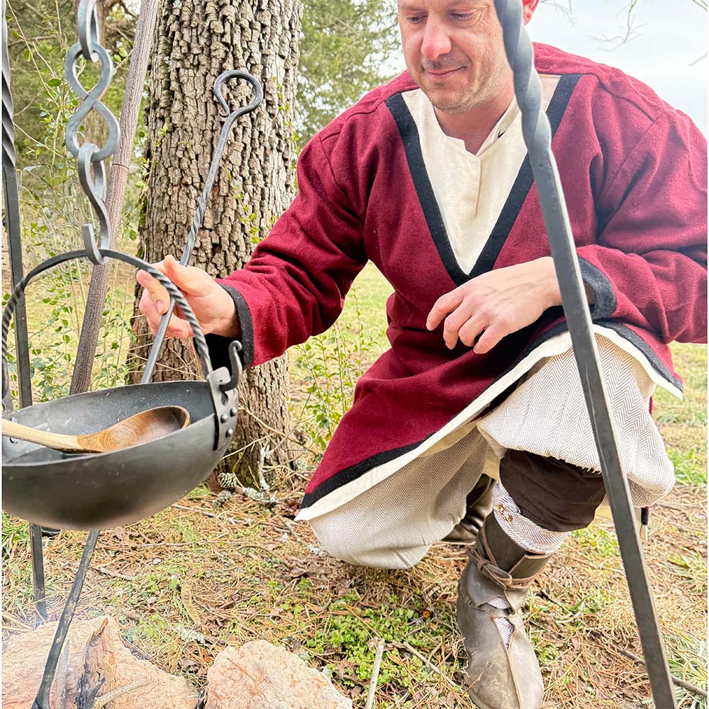 Wearing the Viking Woolen Tunic- Men's, a person tends to a black pot over an outdoor fire pit, with trees and grass in the background.